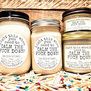 May include: Several soy candles in glass jars with black, silver, and gold lids. Each candle has a label that reads "Light this when you need to CALM THE FUCK DOWN!". The candles are surrounded by matchsticks.