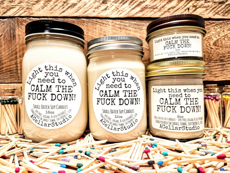 May include: Several soy candles in glass jars with black, silver, and gold lids. Each candle has a label that reads "Light this when you need to CALM THE FUCK DOWN!". The candles are surrounded by matchsticks.