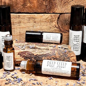 May include: A set of six amber glass roller bottles with black caps and white labels that read "Deep Sleep 100% Natural". The bottles are arranged on a wooden surface with dried lavender flowers scattered around them.