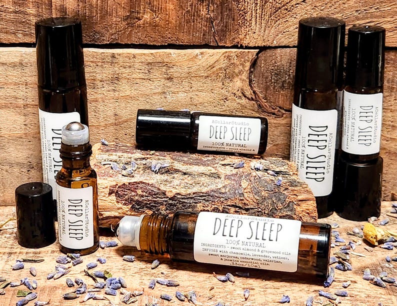 May include: A set of six amber glass roller bottles with black caps and white labels that read "Deep Sleep 100% Natural". The bottles are arranged on a wooden surface with dried lavender flowers scattered around them.
