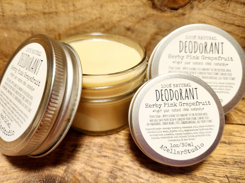 May include: A silver jar with a clear glass lid filled with a yellow-colored natural deodorant. The jar has a label that reads "100% Natural Deodorant Herby Pink Grapefruit *Fight your natural stink naturally*" with a list of ingredients and directions for use. The label also includes the brand name "A Cellar Studio" and the size "1 oz/30 ml".