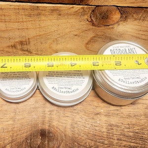 May include: Three silver tins of natural deodorant with labels. The smallest tin is labeled "Herby Pink Grapefruit" and is 1 oz/30 ml. The middle tin is labeled "2 oz/60 ml" and the largest tin is labeled "100% Natural Deodorant 4 oz/120 ml". All three tins are labeled "ACellarStudio".