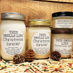 以下が含まれることがあります： ソイワックスキャンドルが入ったガラス瓶が3つ。瓶には「This smells like Christmas break!」とラベルが貼られており、サイズはそれぞれ異なります。瓶は、松ぼっくりとマッチが置かれた木製の表面の上に置かれています。