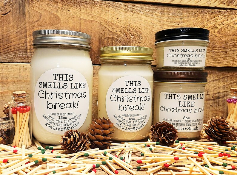 以下が含まれることがあります： ソイワックスキャンドルが入ったガラス瓶が3つ。瓶には「This smells like Christmas break!」とラベルが貼られており、サイズはそれぞれ異なります。瓶は、松ぼっくりとマッチが置かれた木製の表面の上に置かれています。