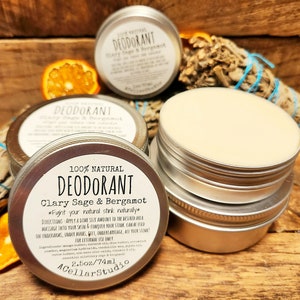 May include: A silver tin with a white label that reads "100% Natural Deodorant Clary Sage & Bergamot *Fight your natural stink naturally*" with directions for use. The tin is open and the deodorant is visible. The tin is sitting on a wooden surface with a piece of dried sage and an orange.