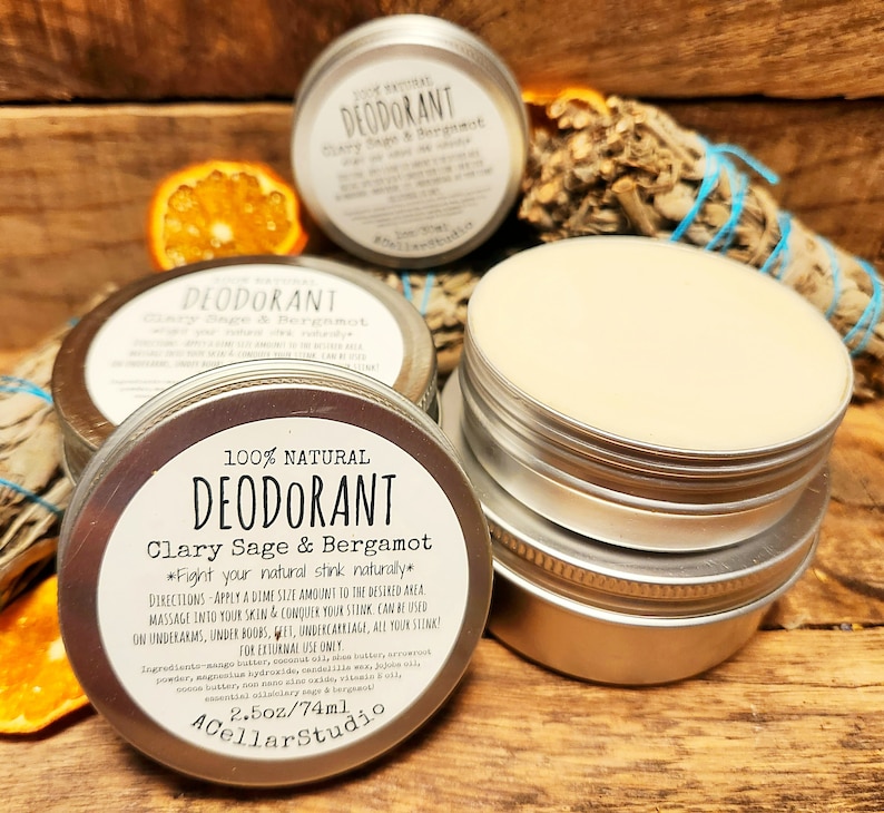 May include: A silver tin with a white label that reads "100% Natural Deodorant Clary Sage & Bergamot *Fight your natural stink naturally*" with directions for use. The tin is open and the deodorant is visible. The tin is sitting on a wooden surface with a piece of dried sage and an orange.