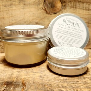 May include: Two silver metal jars of natural deodorant, one larger than the other. The larger jar is labeled "100% Natural Deodorant Herby Pink Grapefruit" and "2 oz/60 ml".