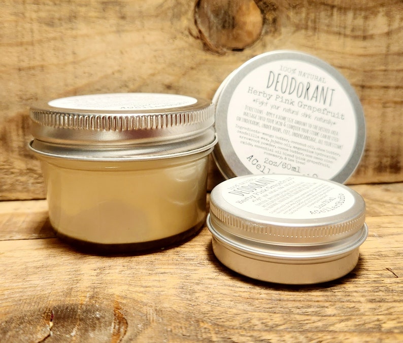 May include: Two silver metal jars of natural deodorant, one larger than the other. The larger jar is labeled "100% Natural Deodorant Herby Pink Grapefruit" and "2 oz/60 ml".