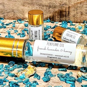 May include: Three glass bottles of perfume oil with gold lids. The bottles are labeled with the text "PERFUME OIL" and the scents "French Lavender & Honey", "Bahama Dreams", and "yurkling clas". The bottles are sitting on a surface covered in blue and gold glitter.