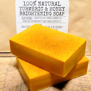 May include: Two bars of bright yellow turmeric and honey soap. The soap is labeled "100% Natural Turmeric & Honey Brightening Soap" and lists the ingredients: Shea Butter Soap, Turmeric, Turmeric Oil, Orange Blossom Raw Honey & Sweet Orange Essential Oil. The label also states "Small Batch, Vegan, & Eco Friendly".