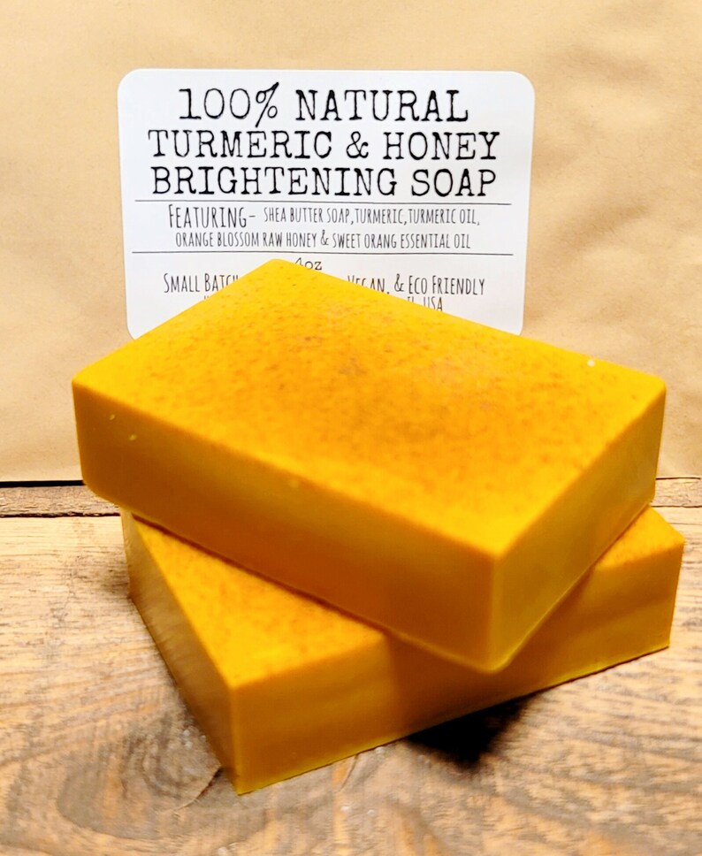 May include: Two bars of bright yellow turmeric and honey soap. The soap is labeled "100% Natural Turmeric & Honey Brightening Soap" and lists the ingredients: Shea Butter Soap, Turmeric, Turmeric Oil, Orange Blossom Raw Honey & Sweet Orange Essential Oil. The label also states "Small Batch, Vegan, & Eco Friendly".