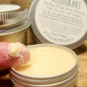 May include: A close-up of a small, silver tin with a white label that reads "100% Natural Deodorant, Herby Pink Grapefruit, with a very citrusy scent". The label also includes the text "2oz/60ml A Cellar Studio". A finger is dipping into the tin, scooping out a small amount of the light yellow, creamy deodorant.