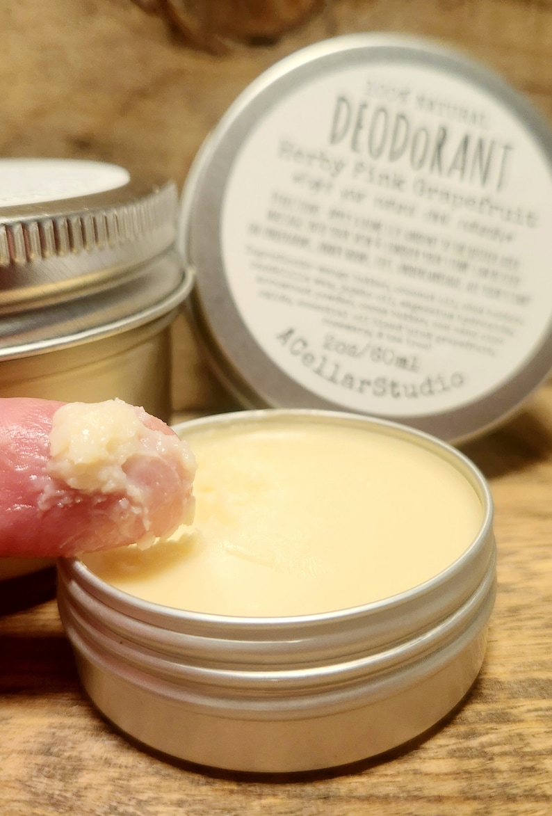 May include: A close-up of a small, silver tin with a white label that reads "100% Natural Deodorant, Herby Pink Grapefruit, with a very citrusy scent". The label also includes the text "2oz/60ml A Cellar Studio". A finger is dipping into the tin, scooping out a small amount of the light yellow, creamy deodorant.