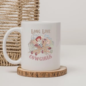 May include: White ceramic mug featuring a cartoon cowgirl riding a horse, with the words "LONG LIVE COWGIRLS" in a vintage font. The design includes a desert landscape with cacti and a setting sun. The mug has a curved handle and sits on a wooden coaster.