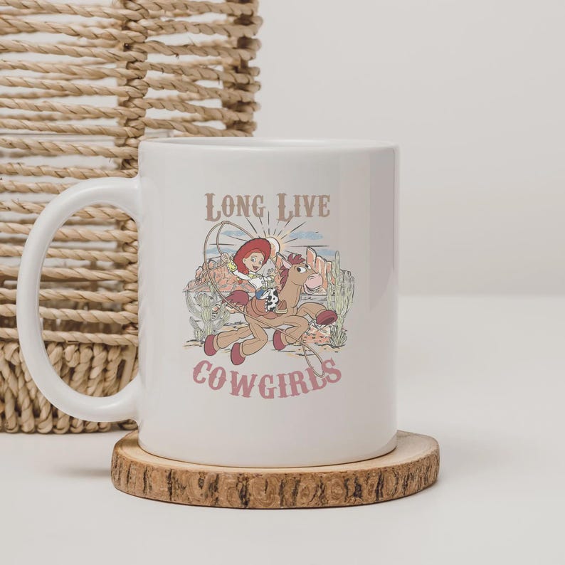 May include: White ceramic mug featuring a cartoon cowgirl riding a horse, with the words "LONG LIVE COWGIRLS" in a vintage font. The design includes a desert landscape with cacti and a setting sun. The mug has a curved handle and sits on a wooden coaster.