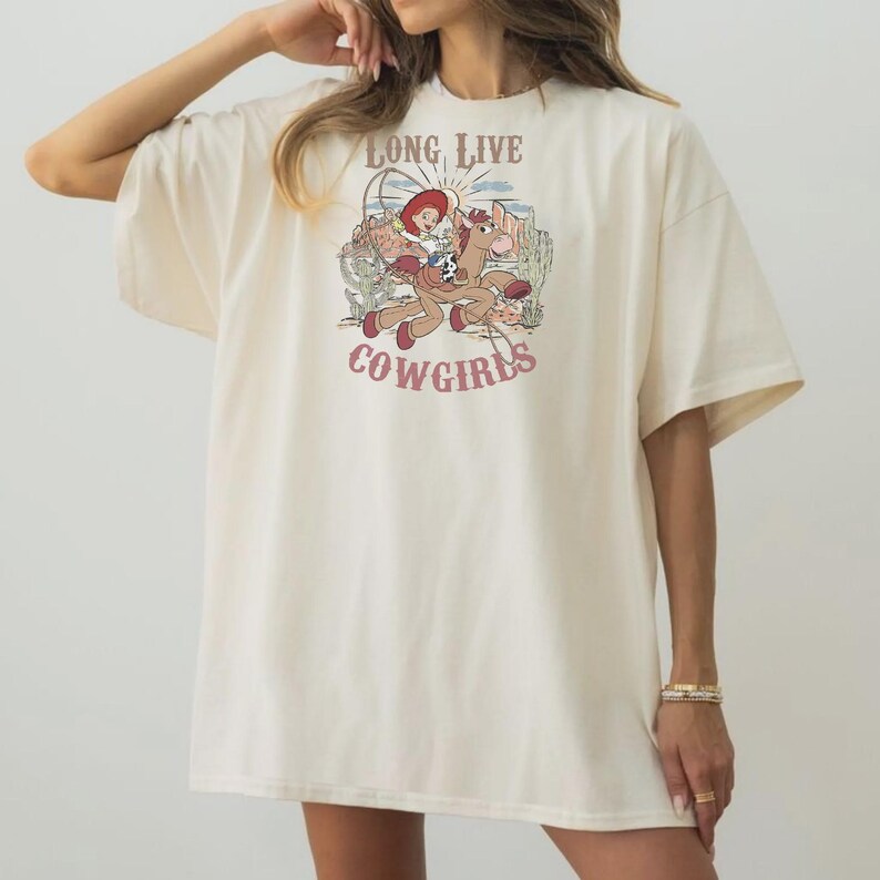 May include: Cream-colored t-shirt featuring a cartoon cowgirl riding a horse, with the text "LONG LIVE COWGIRLS" in a western-style font. The graphic includes desert scenery and a sun. The shirt has short sleeves and a relaxed fit.