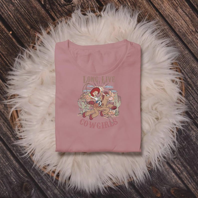 May include: A dusty rose t-shirt featuring a graphic of a cowgirl riding a horse, with the text "Long Live Cowgirls." The design includes cacti and a desert landscape. The shirt is folded on a white fluffy surface.