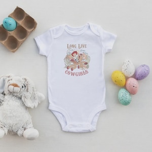 May include: White baby onesie with a graphic of a cowgirl riding a horse, with the text "LONG LIVE COWGIRLS" in red. The onesie is surrounded by Easter eggs and a stuffed bunny.