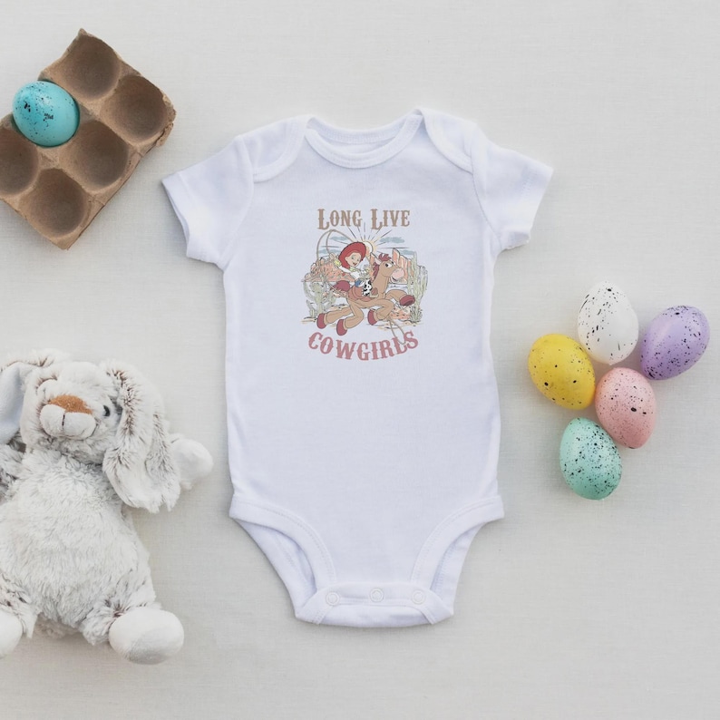 May include: White baby onesie with a graphic of a cowgirl riding a horse, with the text "LONG LIVE COWGIRLS" in red. The onesie is surrounded by Easter eggs and a stuffed bunny.