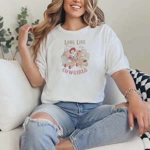 May include: A white t-shirt with the text "LONG LIVE COWGIRLS" and a graphic of a cowgirl riding a horse. The shirt is worn with blue jeans. The overall theme is western and country.