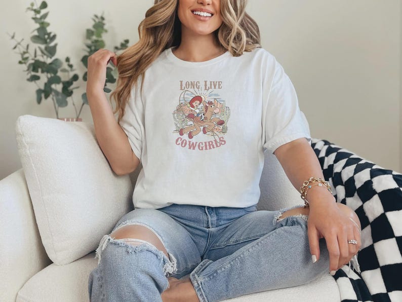 May include: A white t-shirt with the text "LONG LIVE COWGIRLS" and a graphic of a cowgirl riding a horse. The shirt is worn with blue jeans. The overall theme is western and country.