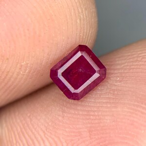 May include: A loose, rectangular-cut ruby gemstone with a deep, saturated red color and faceted edges. The stone displays a clear, polished surface with visible internal clarity and sharp, geometric lines defining its rectangular shape.