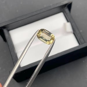 Puede incluir: Una piedra preciosa de color amarillo pálido con corte tipo cojín, sostenida por unas pinzas de metal plateado. La piedra transparente presenta una superficie facetada que refleja la luz, mostrando un tono dorado claro y nítido en toda su forma.