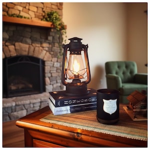 May include: A rustic, black lantern with a lit bulb sits atop two books on a wooden table. A black candle with a label and a small wooden house are also on the table. A stone fireplace and green armchair are in the background.