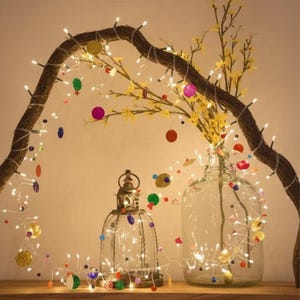 May include: A decorative display featuring a curved branch adorned with string lights and colorful ornaments. A glass lantern and vase hold additional lights and yellow floral accents, creating a warm, inviting ambiance.