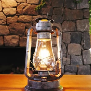 May include: A vintage-style lantern with a clear glass globe and a warm, glowing light. The lantern has a dark metal frame with a handle at the top. It sits on a wooden surface against a stone wall.