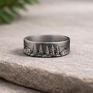 Puede incluir: Un anillo plateado con un diseño de paisaje detallado. El anillo presenta una cordillera y un bosque de árboles. El anillo se muestra sobre una superficie de piedra gris.
