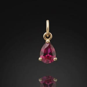 May include: A gold pendant featuring a pear-shaped, deep red gemstone. The gemstone is set in a gold bezel with three prongs. The pendant hangs from a gold loop. The background is black.