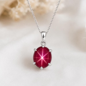 May include: A silver-toned necklace featuring a round, cabochon-cut deep red gemstone with a prominent six-rayed star effect centered on the stone. The pendant is held by a six-prong setting attached to a delicate, thin silver-toned metal chain.