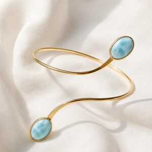 Puede incluir: Una pulsera abierta de tono dorado con un diseño en espiral. Cada extremo de la banda metálica está rematado con una piedra lisa de forma ovalada en color azul claro con patrones de veteado blanco, creando un aspecto simétrico y pulido.