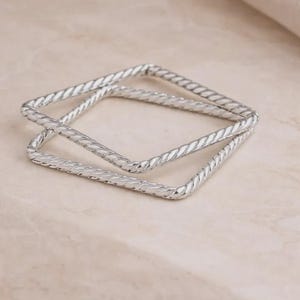 May include: Two silver square bangles with a twisted rope design. The bangles are stacked on top of each other, creating a layered effect. The jewelry is displayed on a light-colored surface.