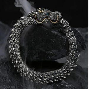 Puede incluir: Pulsera de dragón plateada con detalles de escamas. La cabeza del dragón tiene ojos azules y detalles dorados. La pulsera tiene forma circular, con la cola del dragón unida a su cabeza.