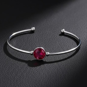 Op de afbeelding: Een zilverkleurige armband met een grote, ronde, rode edelsteen in het midden. De armband heeft een eenvoudig, open ontwerp met afgeronde uiteinden. De edelsteen heeft een diepe, rijke rode kleur en de armband staat tegen een donkere achtergrond.