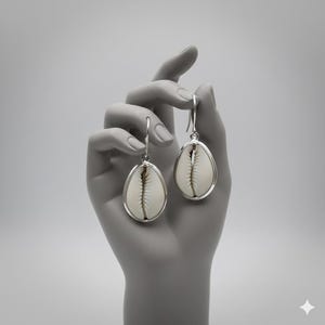 Puede incluir: Un par de pendientes ovalados con una concha blanca enmarcada en plata. Los pendientes cuelgan de ganchos de plata. El fondo es una imagen en escala de grises de una mano sosteniendo los pendientes.