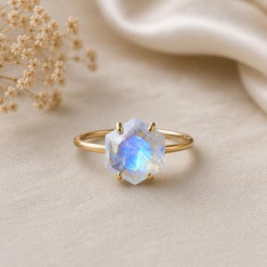 Può includere: Un anello d'oro con una grande pietra di luna esagonale con iridescenza blu e bianca. L'anello ha una sottile fascia color oro, su uno sfondo neutro con illuminazione soffusa. L'anello è un gioiello.