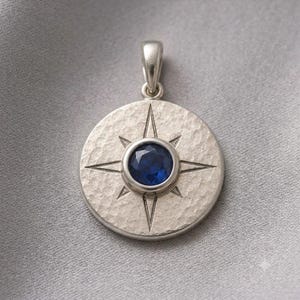 May include: A circular silver pendant featuring a hammered texture and a central faceted deep blue gemstone. The pendant is engraved with a compass rose design consisting of four primary points and four smaller points, topped with a silver bail.