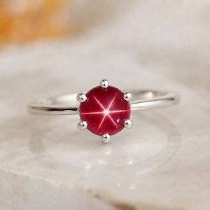 May include: A silver ring featuring a round, deep red star ruby cabochon set in a six-prong mounting. The gemstone displays a distinct six-rayed star effect centered on its surface, held securely by polished metal prongs on a smooth band.