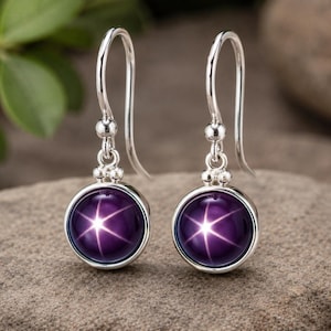 Può includere: Un paio di orecchini in argento con pietre preziose rotonde, viola scuro, con un motivo a stella. Gli orecchini hanno un design a gancio e sono appoggiati su una superficie di pietra grigia.