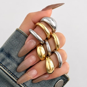 Könnte beinhalten: Eine Sammlung von sieben glatten, polierten Metallringen in abwechselnden Silber- und Goldtönen. Die Ringe zeichnen sich durch ein abgerundetes, gewölbtes Design mit hochglänzender Oberfläche aus. Sie werden gestapelt an mehreren Fingern getragen.