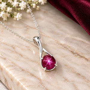 May include: A silver pendant necklace featuring a round, deep magenta star gemstone set in a polished metal frame with a twisted bail design. The pendant hangs from a delicate silver chain, showcasing a prominent six-rayed star light effect.