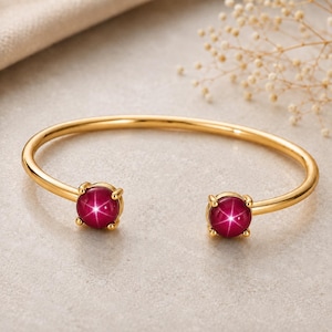 May include: A gold-toned open cuff bracelet featuring two round, deep magenta star-effect cabochon gemstones at each end. The polished metal band is smooth and circular, with each gemstone secured by four small prongs in a classic setting.
