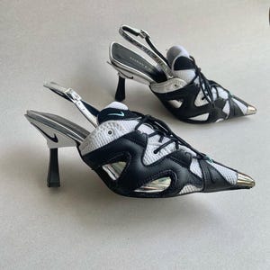 May include: A pair of black, white, and silver heeled shoes with a pointed toe. The shoes feature a mesh and leather design, a black heel, and a silver strap. The Nike logo is visible on the heel.