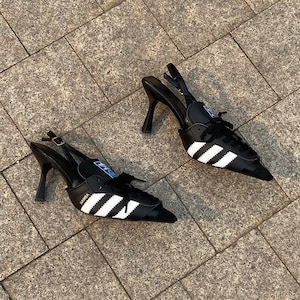 May include: Black high-heeled shoes with a trainer design. The shoes have white stripes, laces, and a pointed toe. The heels are black and curved. The word "SAMBA" is printed on the side of the shoe.
