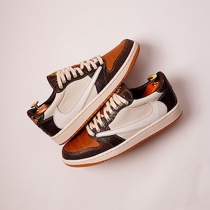 Puede incluir: Dos zapatillas marrones, blancas y negras con cordones blancos. Las zapatillas tienen una puntera marrón, un logotipo blanco y un talón negro. Las suelas son de color marrón claro. Las zapatillas están orientadas hacia el espectador.