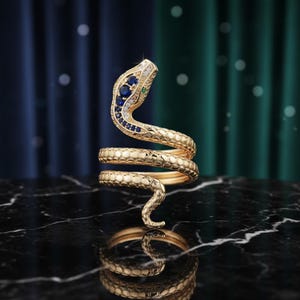 Może przedstawiać: Złoty pierścionek w kształcie węża o spiralnym wzorze. Głowa węża zdobiona jest niebieskimi i białymi kamieniami szlachetnymi oraz zielonym kamieniem. Pierścionek jest prezentowany na czarnej marmurowej powierzchni.