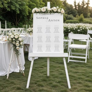 May include: A white easel displaying a wedding seating chart with the names "Sophia & Anton" and table assignments. The easel is decorated with white flowers and greenery. The scene is set outdoors with tables and chairs.